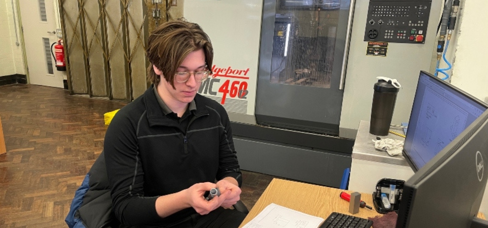 A student working at a desk in a workshop setting, examining a small metal component while seated beside a computer monitor and technical drawings, with industrial machinery visible in the background.